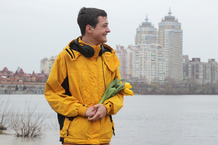 Young handsome teenage boy in yellow coat with ear-phones with a bunch of yellow tulips feeling happy while waiting for a date in rainy day in a big city, outdoor portraitの写真素材