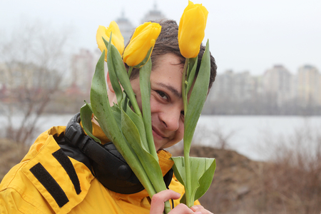 Young handsome teenage boy in yellow coat with ear-phones looking joyful through a bunch of yellow tulips and feeling happy in rainy day in a big city, outdoor portraitの写真素材
