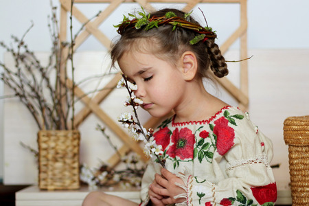Cute girl in traditional embroidered dress and willow wreath stays smelling blooming brunch with closed eyes, dreamily look, Ukrainian history and culture, happy family holiday, closeup indoor portraitの写真素材