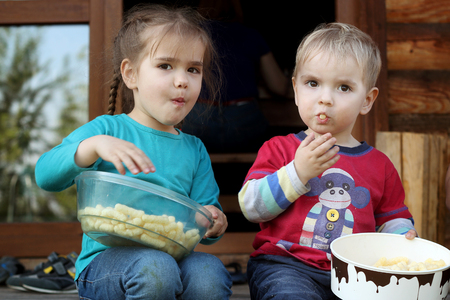 Cute preschool girl and her small brother eating dry corn stick on the wooden stairs near the country house, outdoor close-up, happy family conceptの写真素材