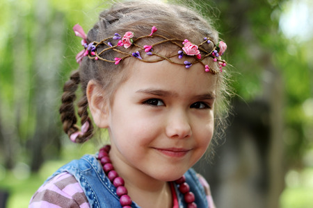 Close-up portrait of little beautiful blonde girl wearing an elegant wreath of leaves and flowers posing in a green meadow. Fashionable attractive child, gorgeous fair hair female, outdoor summer shotの写真素材