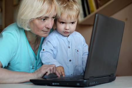 Serious handsome toddler boy together with his grandmother working on the laptop computer at home,...