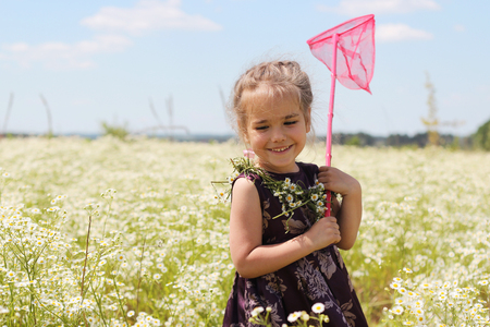 Cute funny little girl with wildflower wreath on her neck and butterfly net staying in chamomile field, beauty and health care, happy childhood, active summer vacation, close-up outdoor portraitの写真素材