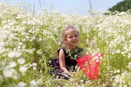 Cute funny little girl with wildflower wreath on her neck and butterfly net sitting tired in chamomile field, beauty and health care, happy childhood, active summer vacation, close-up outdoor portraitの写真素材