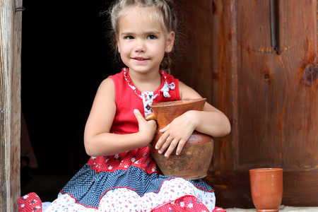 Attractive young girl sitting on the threshold of countryside house and holding an earthenware pot with organic milk, summer outdoor, healthy lifestyle conceptの写真素材