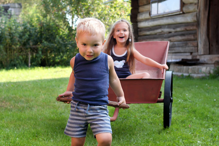 Smiling siblings, sister and brother, playing together in the backyard during their summer vacation, they riding each other in the barrow, happy childhood concept, summer outdoorの写真素材