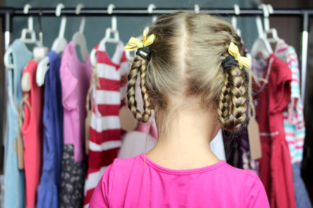 Cute preschooler girl choosing a new dress among a lot of clothes in the child fashion store; back view. Sale, gifts, Christmas, holidays; beauty and fashion concept. Indoor portraitの写真素材
