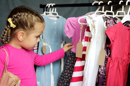 Happy cute preschooler girl choosing a new dress among a lot of clothes in the child fashion store; she is surprised. Sale, gifts, Christmas, holidays; beauty and fashion concept. Indoor portraitの写真素材