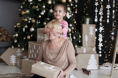 Pretty little girl closing eyes to her grandmother to give a Christmas gift before New Year Tree, merry Christmas concept, happy family, lifestyle indoor, studio shot の写真素材