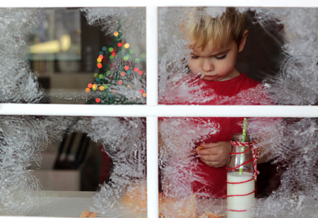 Dreaming little toddler boy in red t-shirt waiting for Santa near frosty window, blurred lights of Christmas Tree, winter holiday concept, indoor portrait  の写真素材