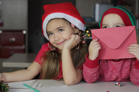 Cute small siblings, boy and girl in red hat, writing a letter to Santa Claus and thinking about his presents for them, Christmas background, winter holiday conceptの写真素材