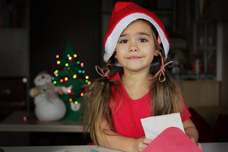 Funny small girl in red hat holding a letter to Santa Claus, blurred decorated Christmas Tree on background, family holiday concept, indoor portraitの写真素材