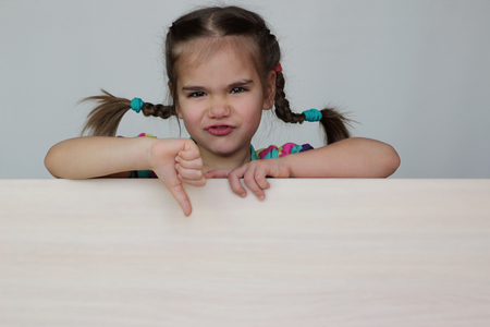 Cute 5 years old girl with funny pigtails showing dislike gesture with thumb down on white board, space for copy, advertising and announcement concept, studio shot over white backgroundの写真素材