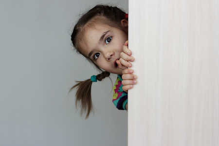 Cute 5 years old girl with funny pigtails peeping out through the blank wall over white background, space for copy, advertising and announcement concept, studio shotの写真素材