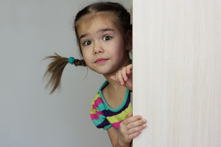 Cute 5 years old girl with funny pigtails peeping out through the blank wall over white background, space for copy, advertising and announcement concept, studio shotの写真素材