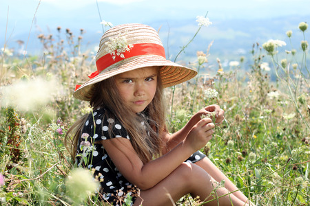 Pretty little girl in an elegant dress and straw hat relaxing on the top of the mountains, summer, vacation, travel concept, she gathers flowers, outdoor portraitの写真素材