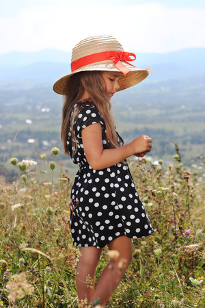 Pretty little girl in an elegant dress and straw hat relaxing on the top of the mountains, summer, vacation, travel concept, she gathers flowers, outdoor portraitの写真素材