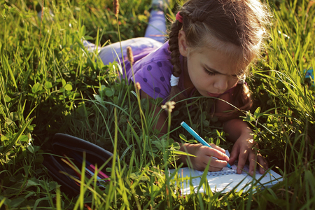 Pretty 5-6 years old girl lying on the green grass and drawing with colorful pencils in the mountains during sunset, beautiful summer evening, family vacation conceptの写真素材