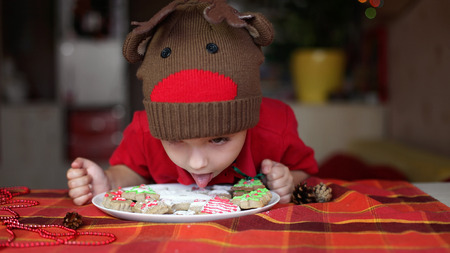 Cute toddler boy in the hat of reindeer eating a cookie from the festive plate with Christmas sweets, classical red and brown Christmas color, winter holidays conceptの写真素材