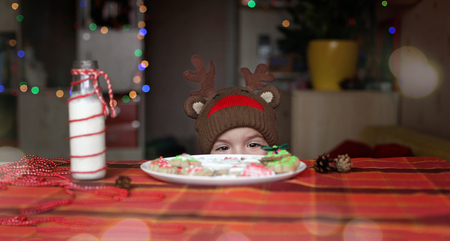 Cute toddler boy in the hat of reindeer stretching his hand to festive Christmas plate with sweet cookies, classical red and brown Christmas color, winter holidays conceptの写真素材