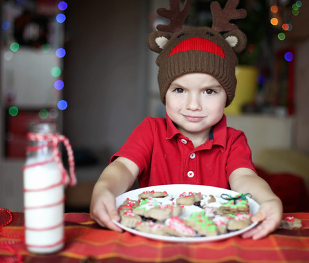 Cute toddler boy in the hat of reindeer eating a cookie from the festive plate with Christmas sweets, classical red and brown Christmas color, winter holidays conceptの写真素材