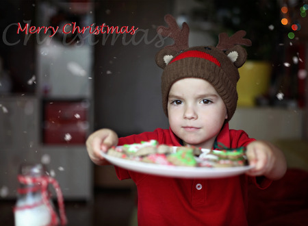Cute toddler boy in the hat of reindeer eating a cookie from the festive plate with Christmas sweets, classical red and brown Christmas color, winter holidays concept, greeting card formatの写真素材