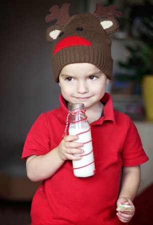 Cute toddler boy in the hat of reindeer drinking milk and eating a cookie from the festive plate with Christmas sweets, classical red and brown Christmas color, winter holidays conceptの写真素材