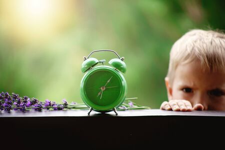 Toddler boy switching off a green alarm clock with lavender flowers on the wooden table of a veranda...