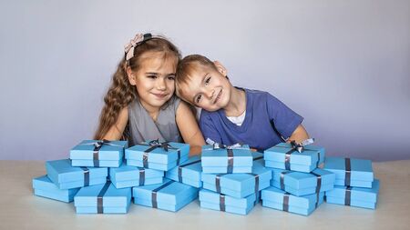 Cute little girl with her smaller brother near a huge pile of blue gift boxes with gray ribbons over white studio backgroundの写真素材