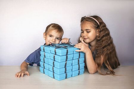 Cute little girl with her smaller brother near a huge pile of blue gift boxes with gray ribbons over...