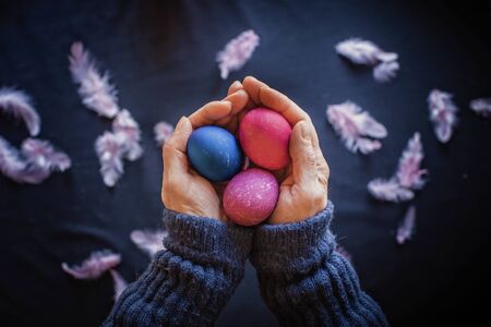 Alternative Easter composition in dark style with classic blue and pink color eggs, feather and hands of senior woman over black background, spring season holidays conceptの写真素材