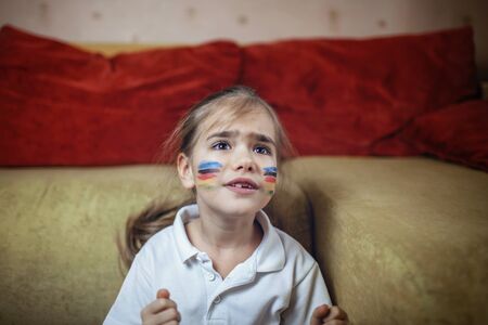 Cute young sport fan with colored cheeks sitting on sofa, watching sport games and cheering for her team on TV at home, real emotions, sports event and fun supporting conceptの写真素材
