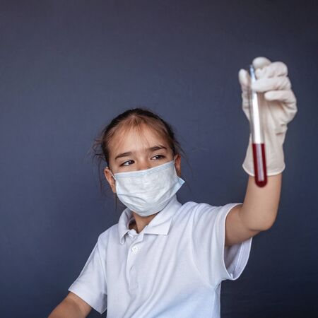 Cute kid wearing respirator mask and medicine gloves playing in a doctor and holding a blood test result for the new rapidly spreading Coronavirus over blue background, health and medicine conceptの写真素材