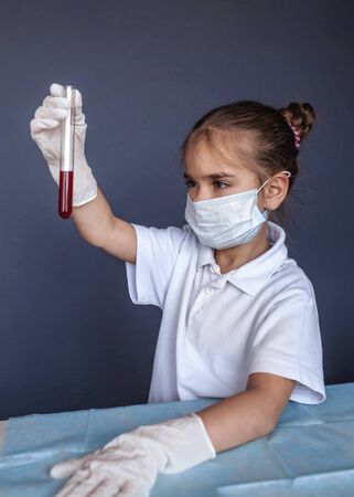 Cute kid wearing respirator mask and medicine gloves playing in a doctor and holding a blood test result for the new rapidly spreading Coronavirus over blue background, health and medicine conceptの写真素材