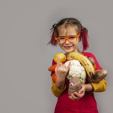 Freakish kid with pinky hair in eyeglasses holds misshapen wrong color fruits and vegetables, giants, with tiny spots and scars, isolated over white background, garbage and waste food trend conceptの写真素材
