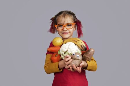 Freakish kid with pinky hair in eyeglasses holds misshapen wrong color fruits and vegetables, giants, with tiny spots and scars, isolated over white background, people and waste food trend conceptの写真素材
