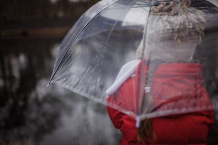 Sad pretty 7-8 years girl in red coat with transparent umbrella walking alone in early spring...