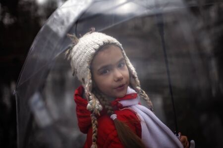 Happy pretty 7-8 years girl in red coat with transparent umbrella walking alone in early spring forest, emotional outdoor portraitの写真素材