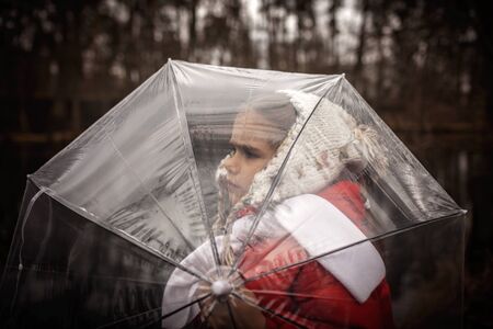 Sad pretty 7-8 years girl in red coat with transparent umbrella walking alone in early spring forest, emotional outdoor portraitの写真素材