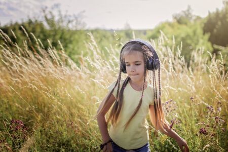 Cool 7-8 years old school girl with many braids and headphones listening music and dancing at sunset among wheat and flower field, music and nature concept, outdoor emotional lifestyle portraitの写真素材