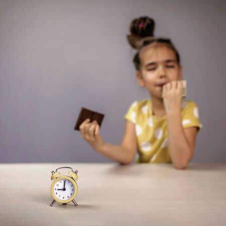 Stylish school girl eating chocolate bars with pleasure and taking no account of clock before her,...