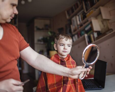 Middle aged father cutting hair to his little son by himself at home looking video broadcast on...