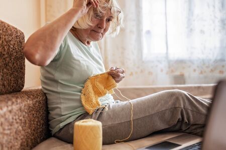 Concentrated old woman sitting on sofa, looking on the laptop screen and knitting a wool sweater at home during quarantine time, online hobby time, stay safe conceptの写真素材