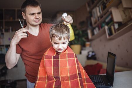 Middle aged father cutting hair to his little son by himself at home looking video broadcast on laptop, hairstyle online class, stay home and life during lockdownの写真素材