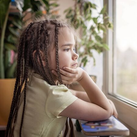 Cute sad little kid sitting on the window sill and looking on the street, dream about outdoor activity during quarantine lockdown, indoor lifestyle, concept of sorrowの写真素材