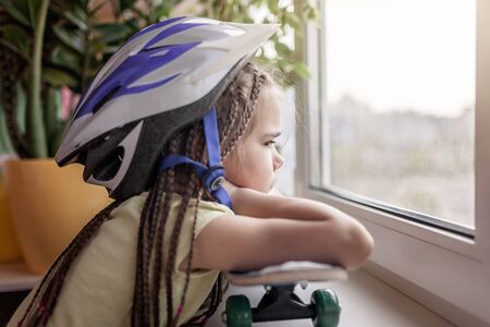 Cute sad little kid sitting on the window sill and looking on the street, dream about outdoor...