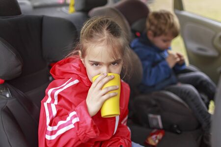Child car safety, family travel. Pretty schoolchild sitting in car seat and holding a yellow cup, lunch during car journey, vacation after lockdownの写真素材