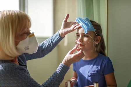 Multigenerational relations, care about seniors in new normal life, grandmother playing in beauty salon and helping grandkid to do makeup, they are wearing in medical respirators, social distance ruleの写真素材