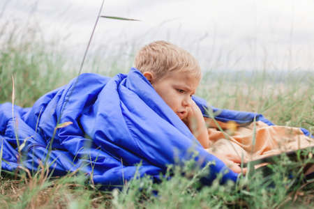 Local vacation. Cute boy having rest and dreaming alone in sleeping bag on the top of the mountain not far from village during local vacation, family summer weekend, social distance lifestyleの写真素材