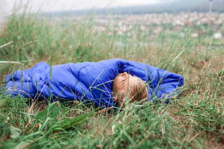Local vacation. Cute boy having rest and dreaming alone in sleeping bag on the top of the mountain not far from village during local vacation, family summer weekend, social distance lifestyleの写真素材